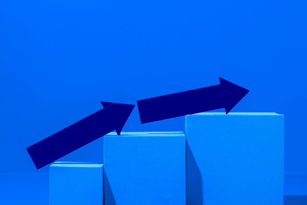 A minimalist blue bar chart with two dark blue arrows pointing upward across rising blocks, symbolizing slowing growth.