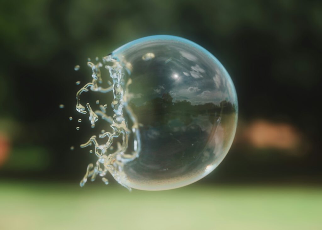 A close-up of a soap bubble mid-burst, with water droplets flying outward and a blurred green background behind it.