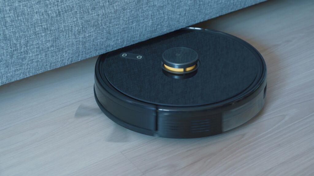 A round black robot vacuum bumps into the base of a gray fabric sofa while cleaning a light wood floor.